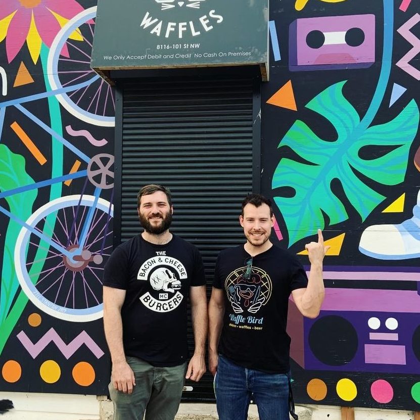 Two men pose and smile in front of a mural-decorated storefront