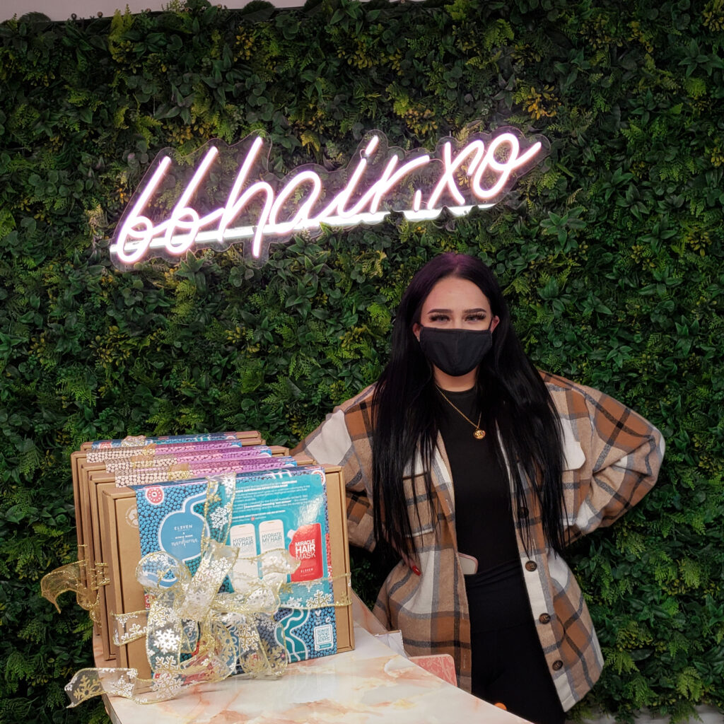 A woman with long black hair and a matching mask smiles under a neon sign with the letters bb hair xo