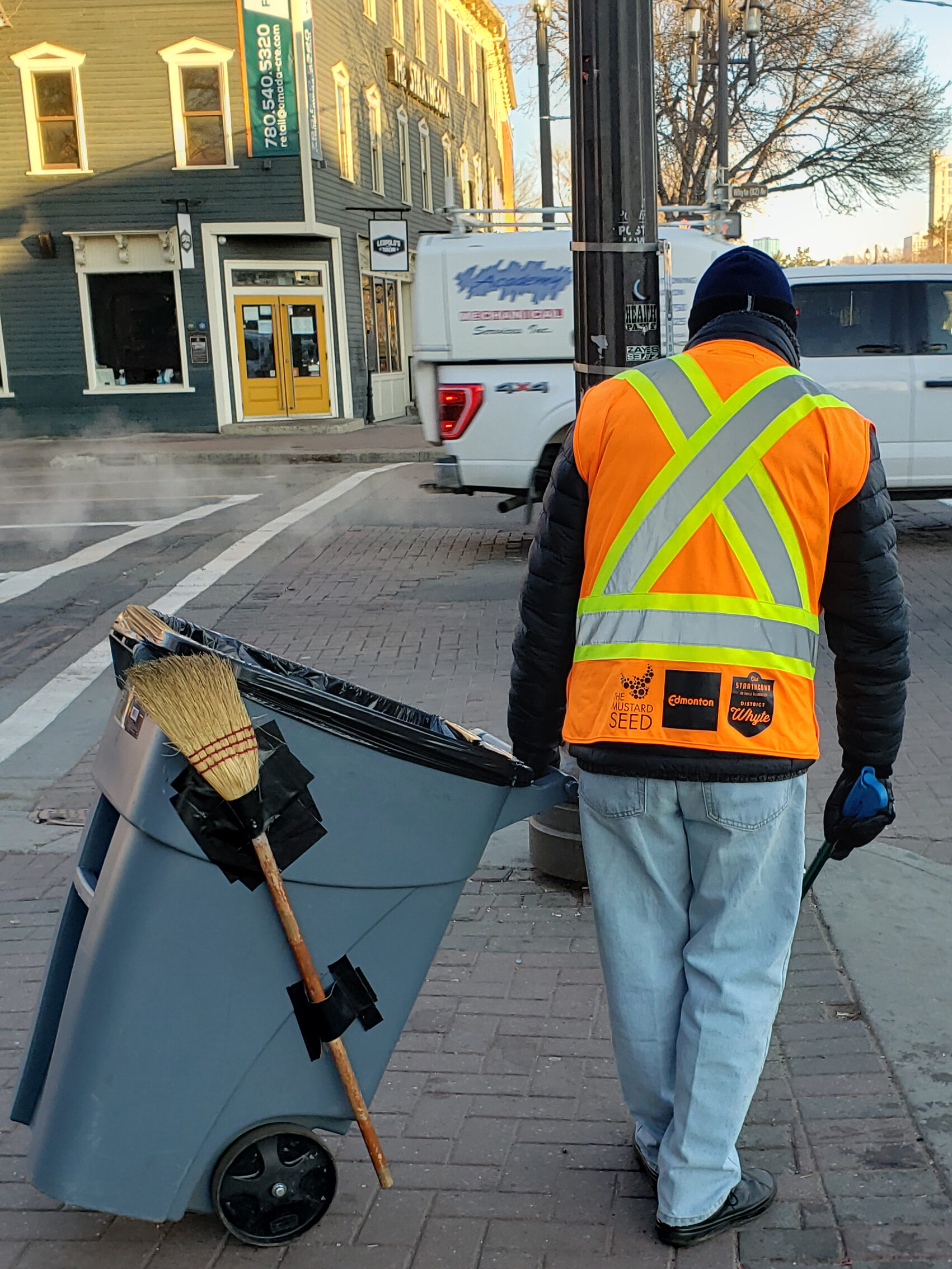 A member of the Old Strathcona Clean Team in an orange vest sweeps Whyte Avenue while pulling a grey bin