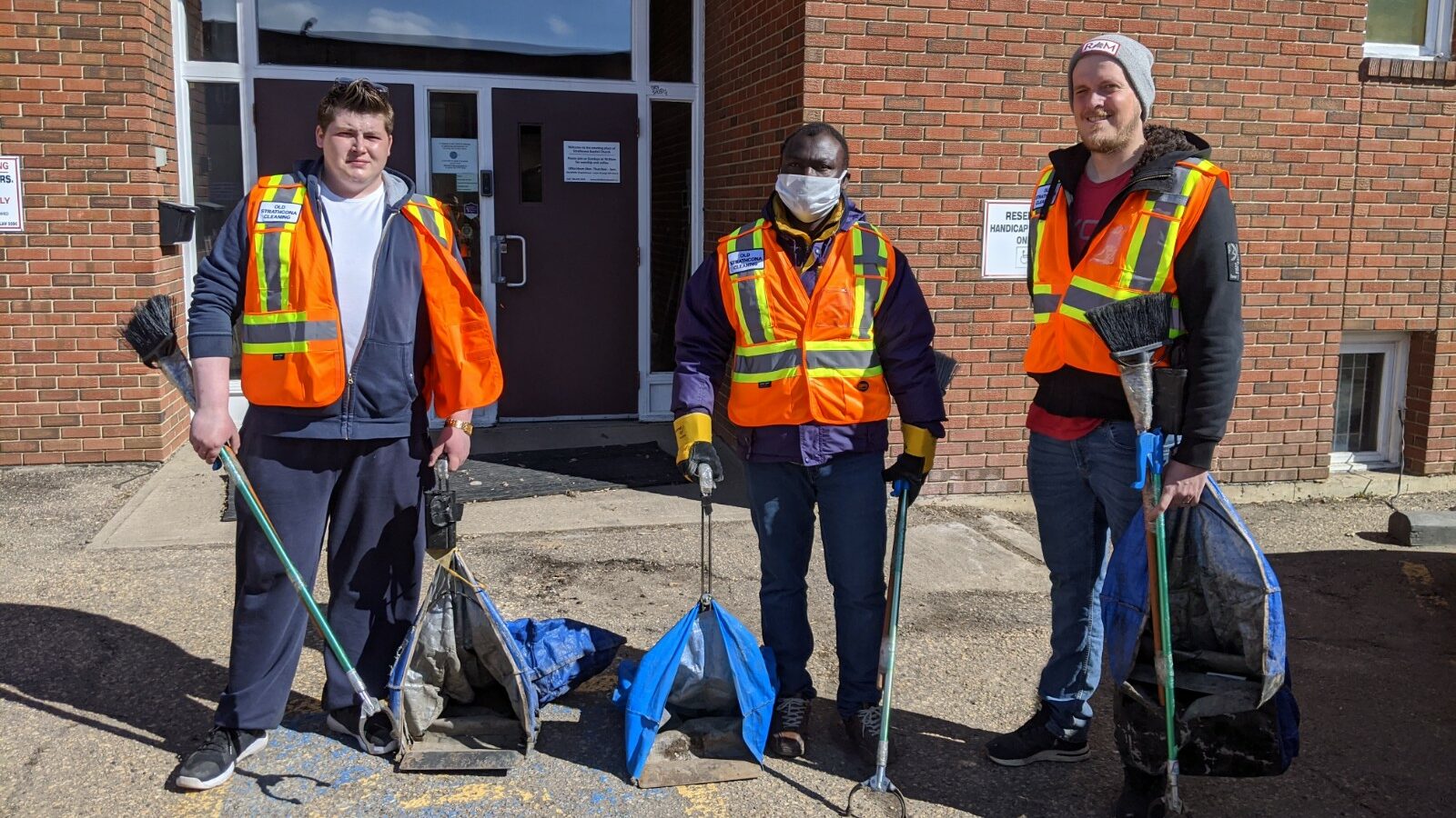 Three members of the Old Strathcona Clean Team pose for a photo wearing orange vests, holding cleaning grabbers and blue bags