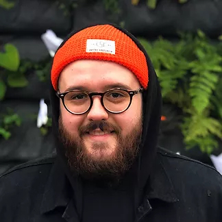 A young bearded man with white skin tone offers a small smile. He's wearing round black-rimmed glasses and an orange toque, standing in front of green plants