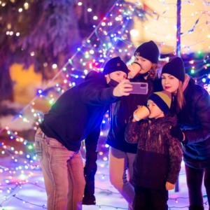 A family smiles and poses for a selfie in front of lit-up trees